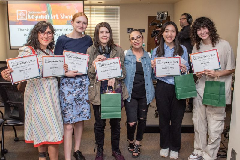 A group of students and art teachers hold up award certificates and prize bags at the Clackamas ESD Regional Art Show awards ceremony