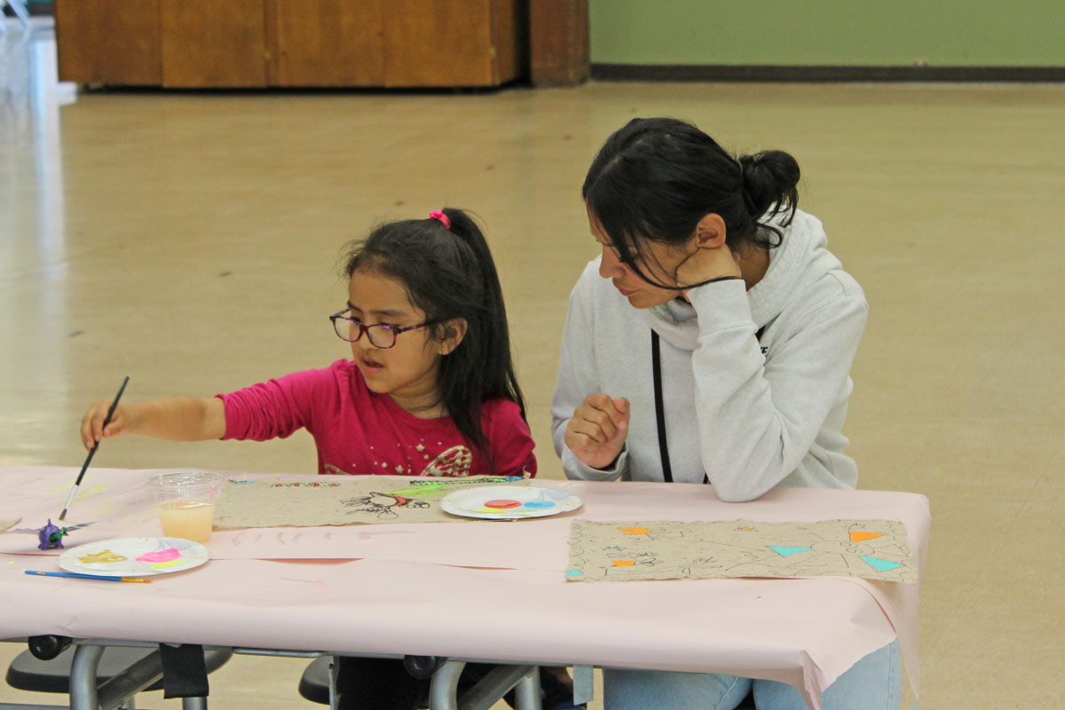 Migrant education program teacher sits beside a student while she is working on an art project
