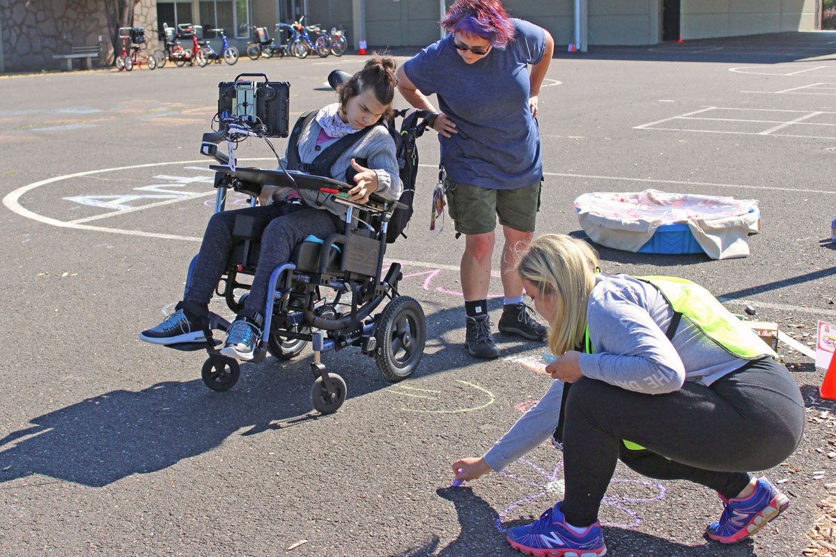 Life Enrichment Education Program student watches as a staff member draws her a flower on the sidewalk with purple chalk
