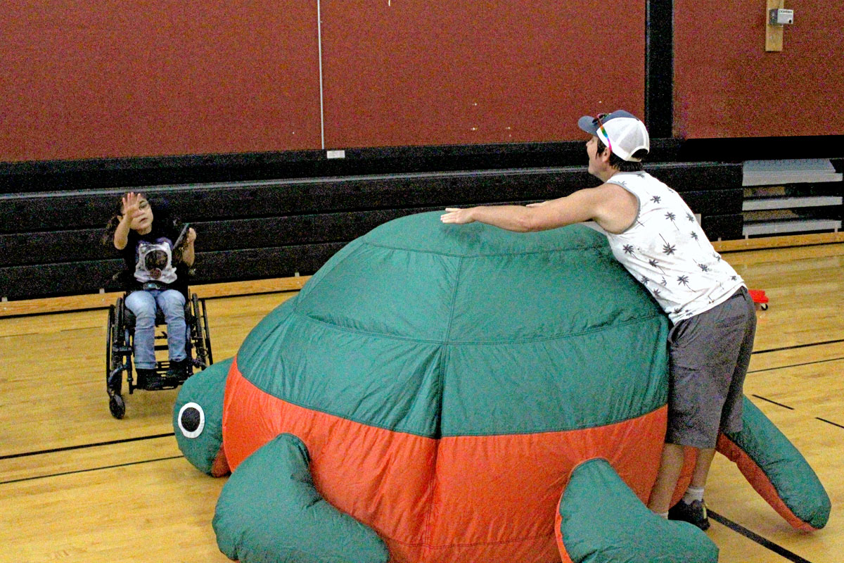 Life Enrichment Education Program staff member plays hide and seek behind a giant inflatable turtle with a LEEP student in the school gym