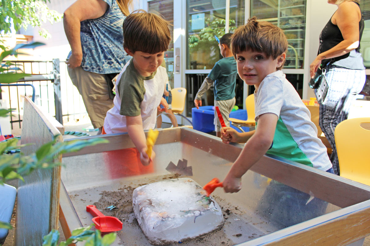 Two preschool children use toy hammers to get dinosaur figurines out of a block of ice