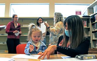 A Clackamas ESD staff member colors with a young girl while other staff members discuss the girl's developmental screening with her mother at a free screening at Gladstone Public Library