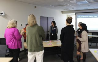 High School Success Network members stand in circle around a table filled with post it notes with a slide reading "Student Focus Group Data" in the background
